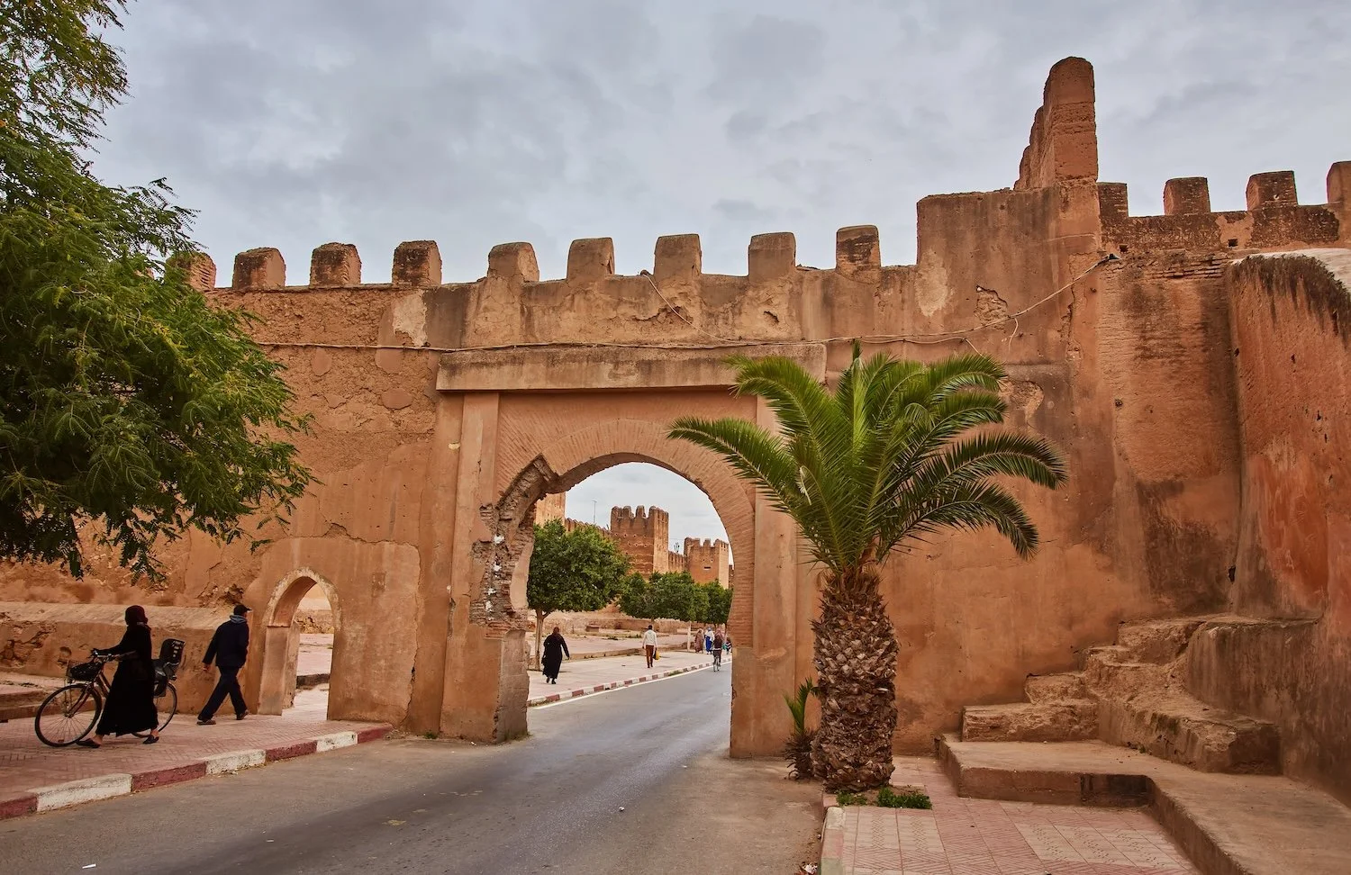 An ancient reddish-brown, fortified wall with crenellations and an arched gateway, situated on a street with pedestrians, a bicycle, a palm tree, and some green trees, under a partly cloudy sky.