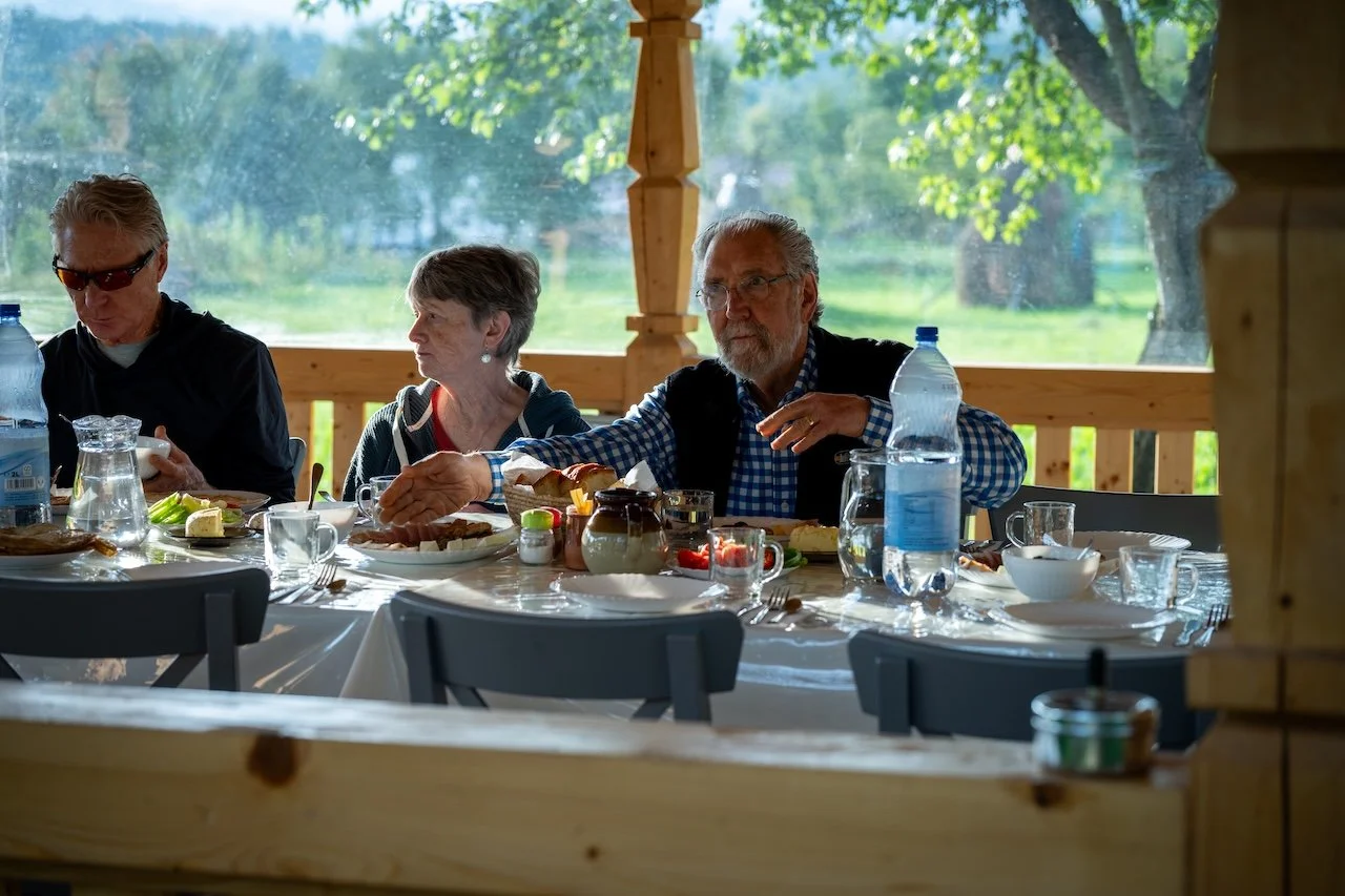 Three older adults sitting at a table having an outdoor meal with a scenic green landscape in the background.