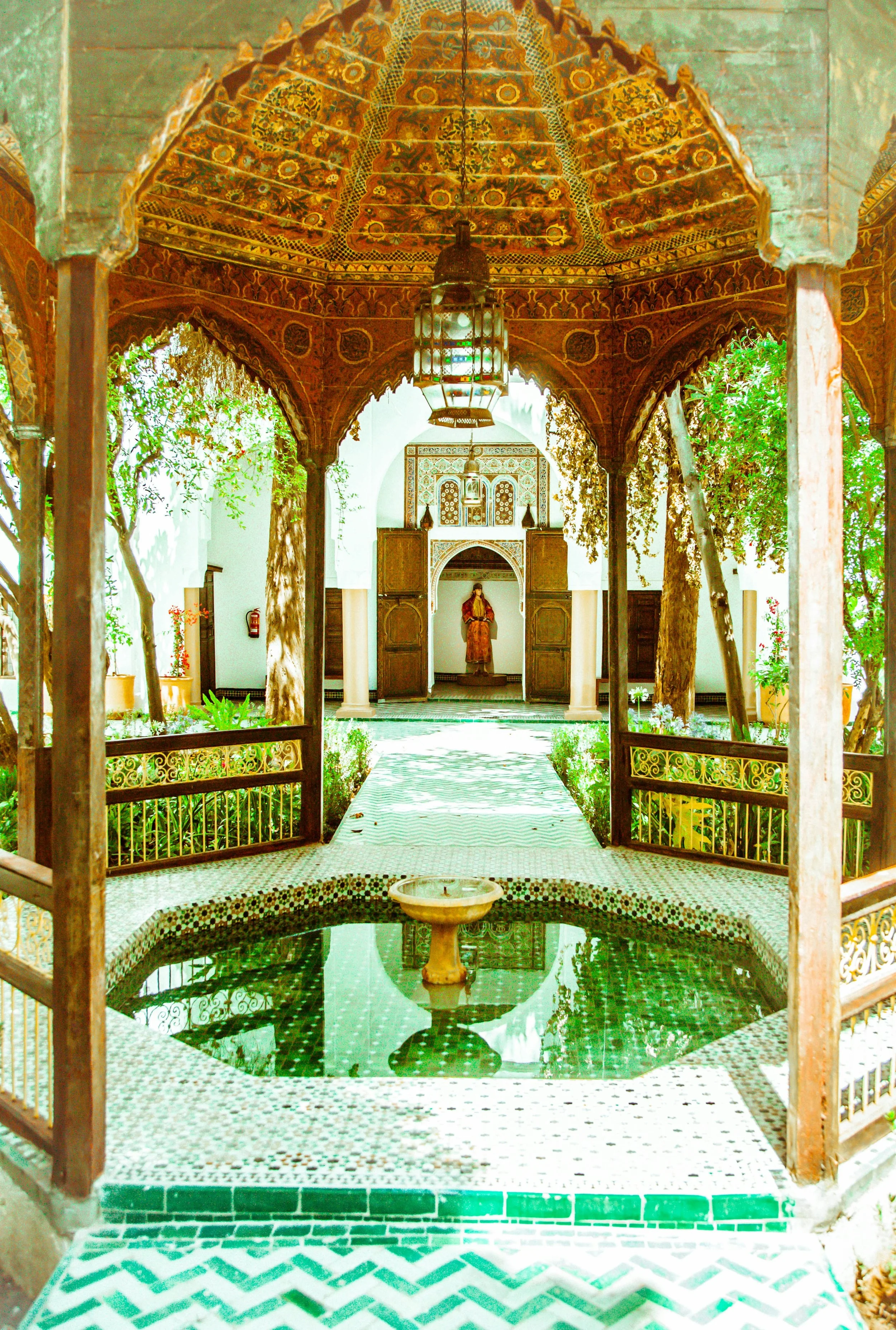 Indoor courtyard with a central fountain, ornate wooden canopy, and archways leading to a statue of a person in traditional attire, surrounded by trees and plants.