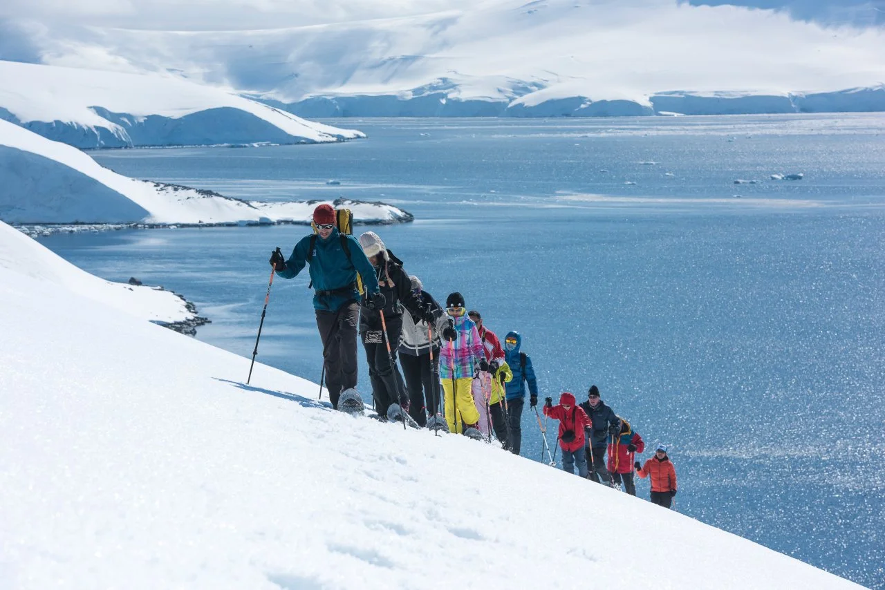 A group of people in colorful winter clothing snowshoeing on a snowy slope near a large body of water with ice and snow-covered mountains in the background.