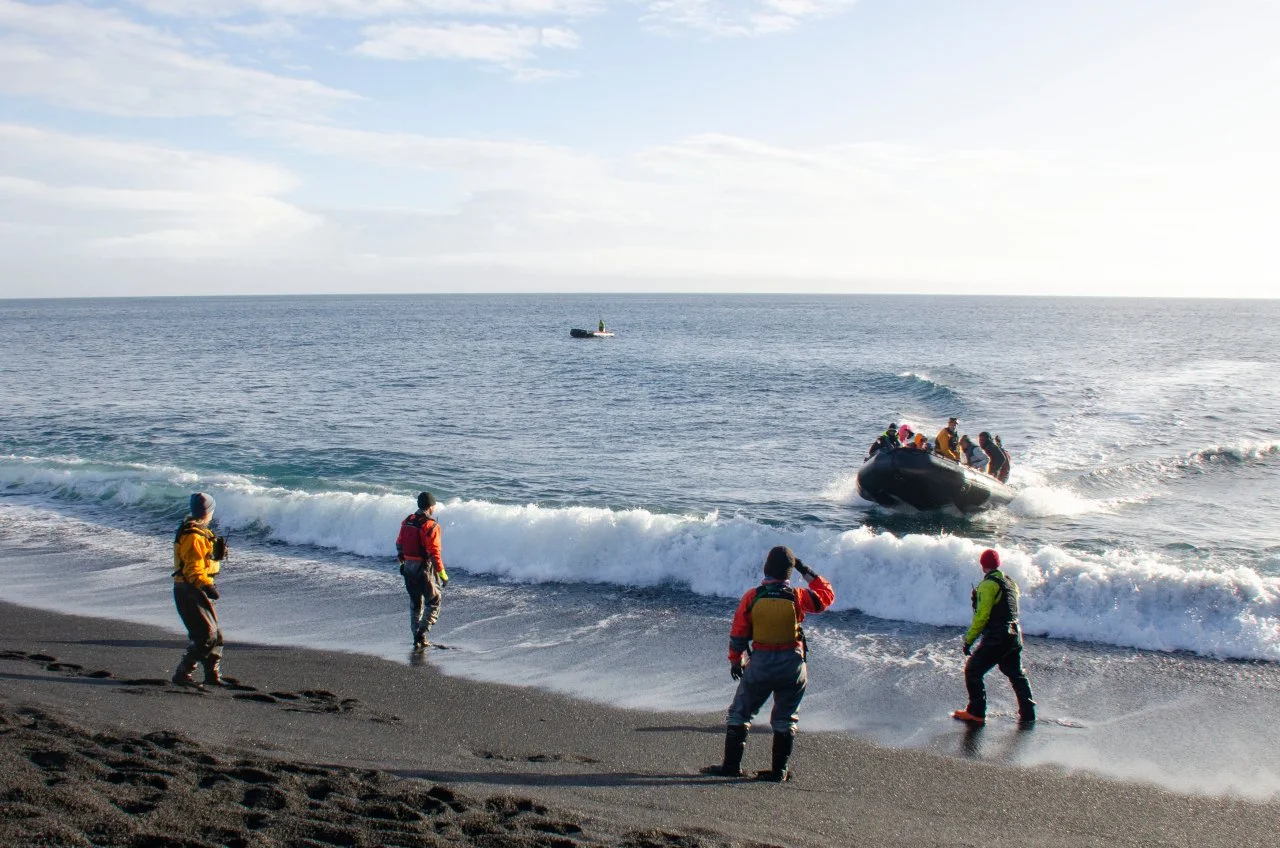 People on a beach watching a small boat with passengers approaching from the water.