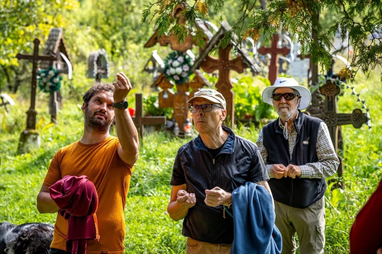 Three men in a cemetery surrounded by crosses and decorated graves, one gesturing while speaking, two wearing hats and sunglasses.