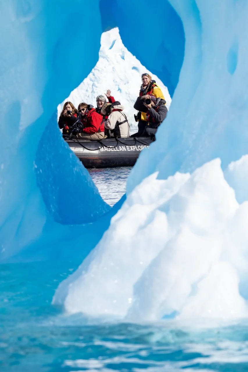 A group of people in winter clothing taking photos from a small boat in icy waters, seen through a hole in a large iceberg with a mountain of snow in the background.