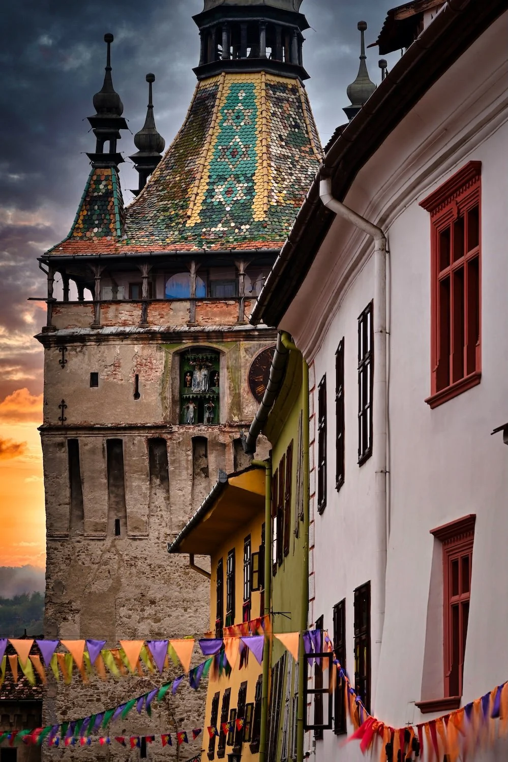 Old fortified tower with a colorful, patterned, tiled roof and a clock, situated next to a row of vibrant, historic buildings decorated with ribbons and flags at sunset.