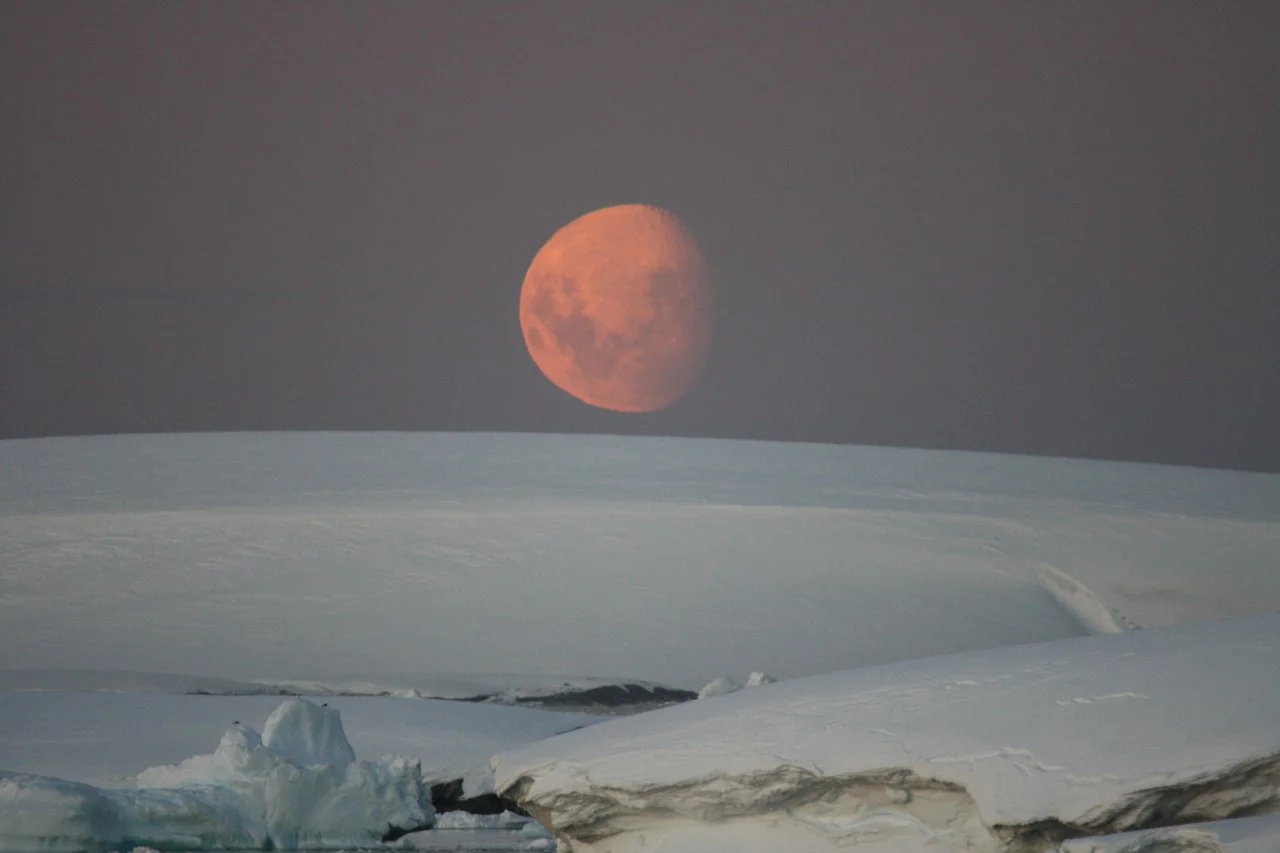 A partially eclipsed orange moon rising over a snowy landscape with ice and snow formations.