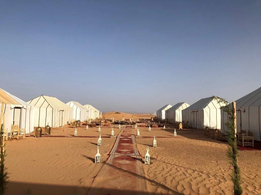A row of white tents in a desert with a sandy path in the center, decorated with lanterns along the walkway and small benches on the sides.