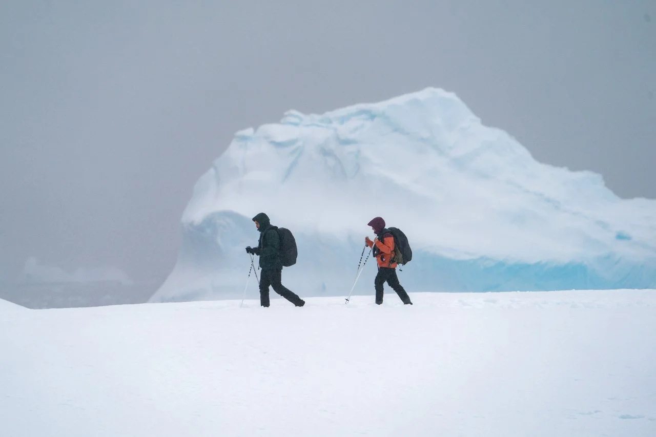 Two hikers in winter gear walking on snow with a large iceberg in the background.