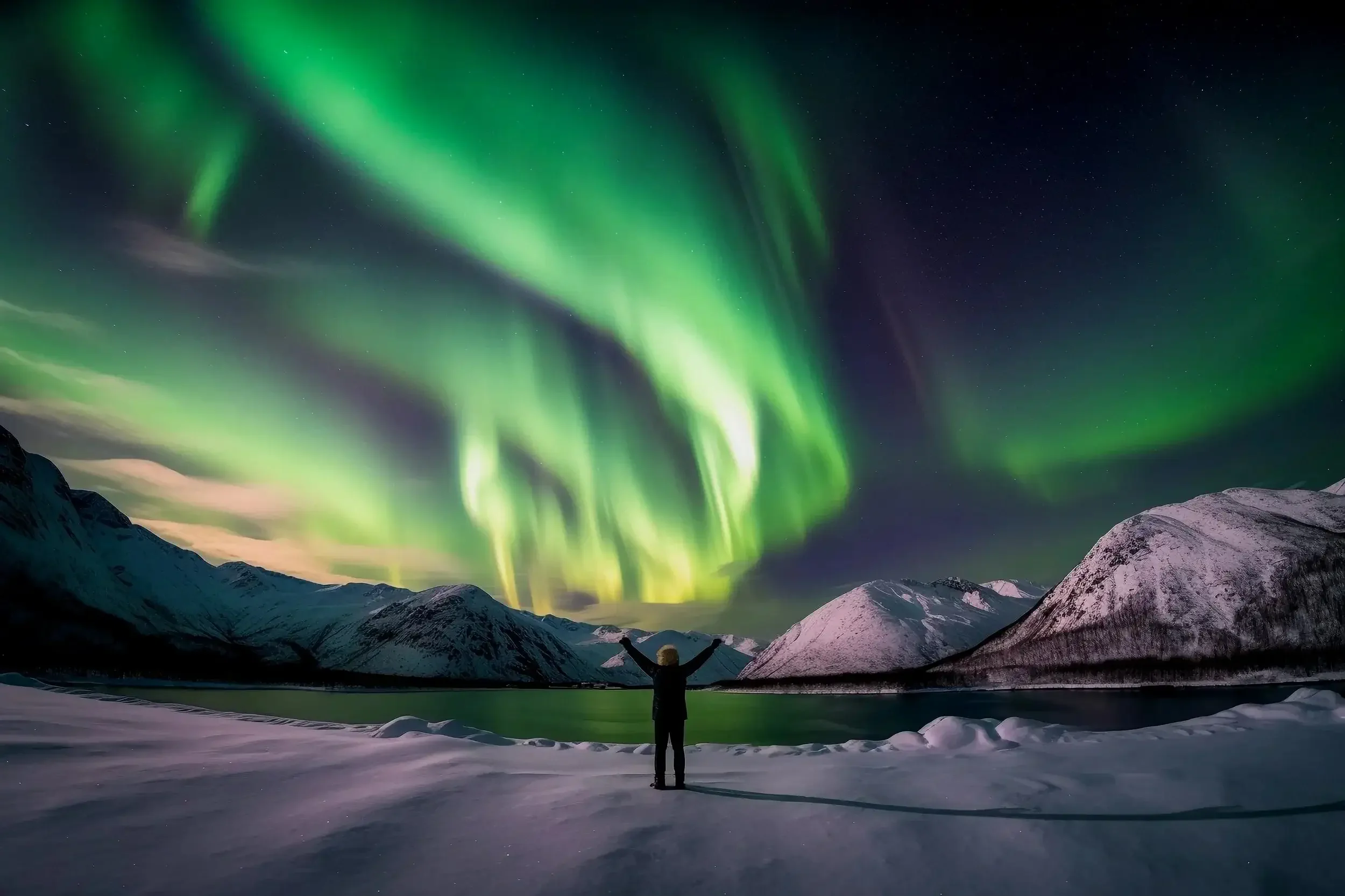 Man standing under northern lights at a lake with snow covered mountatins