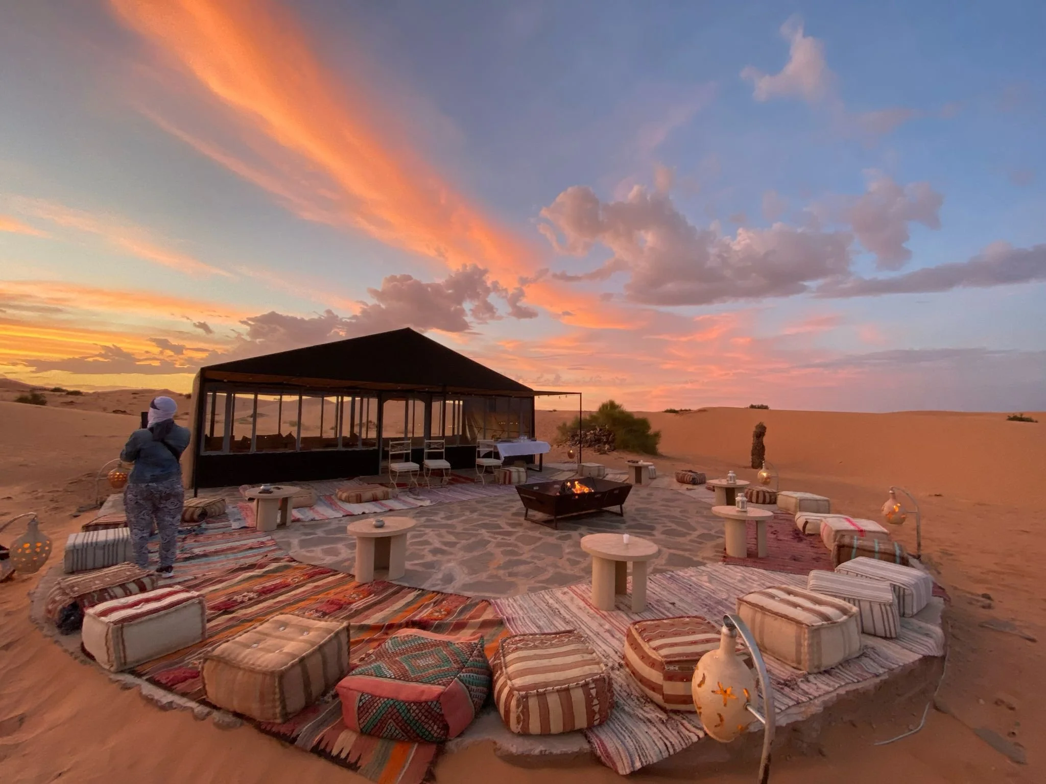 Outdoor desert lounge setup with colorful rugs, cushions, a fire pit, and decorative lanterns during sunset.