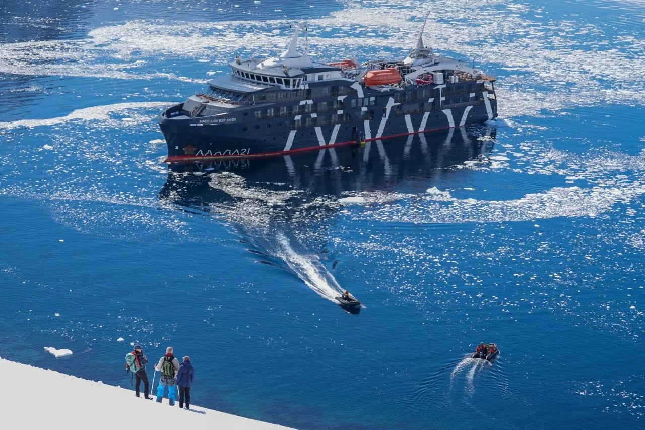 Aerial view of Magellan Discoverer from Antarctic glacier with people on shore and zodiac boats on the water