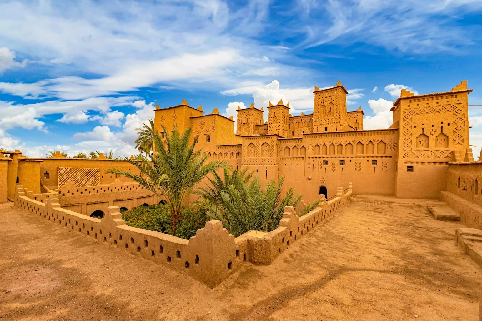 An ancient desert fortress with mud-brick walls and towers, surrounded by a small garden with palm trees under a partly cloudy sky.