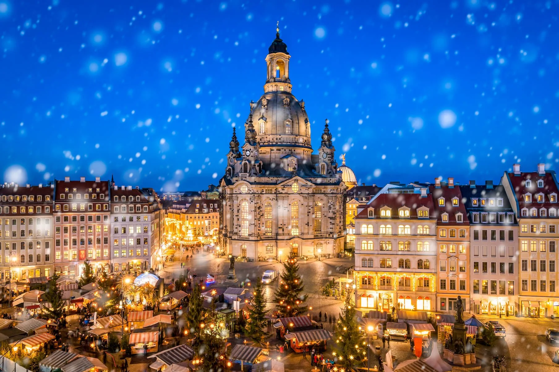 Nighttime view of the Dresden Frauenkirche in Germany with Christmas market stalls and festive lights in the foreground, snow falling across the city