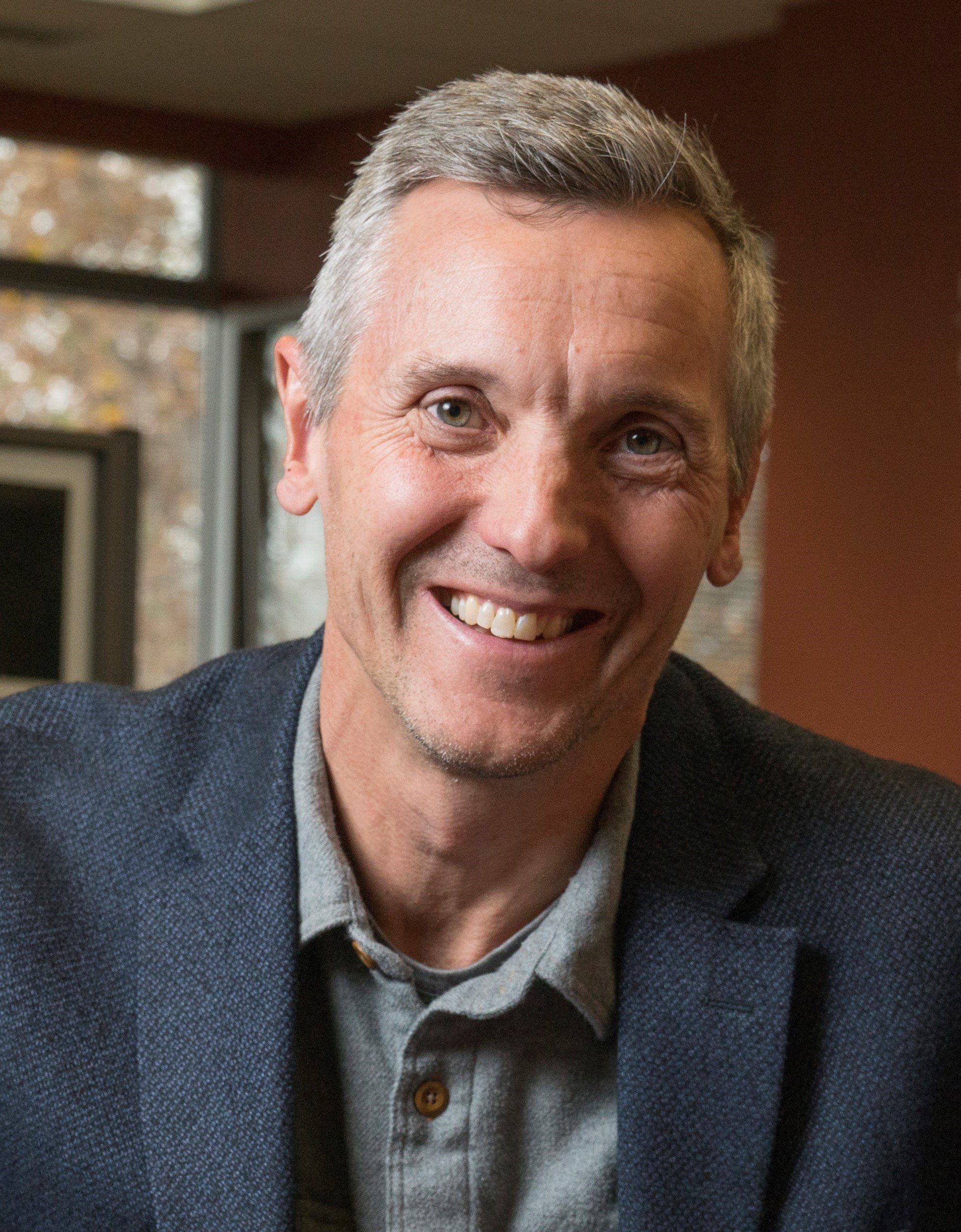 Central America expert Dean McGovern, smiling and wearing a gray button-up shirt and dark blazer, in an indoor setting with large windows and autumn trees visible outside.