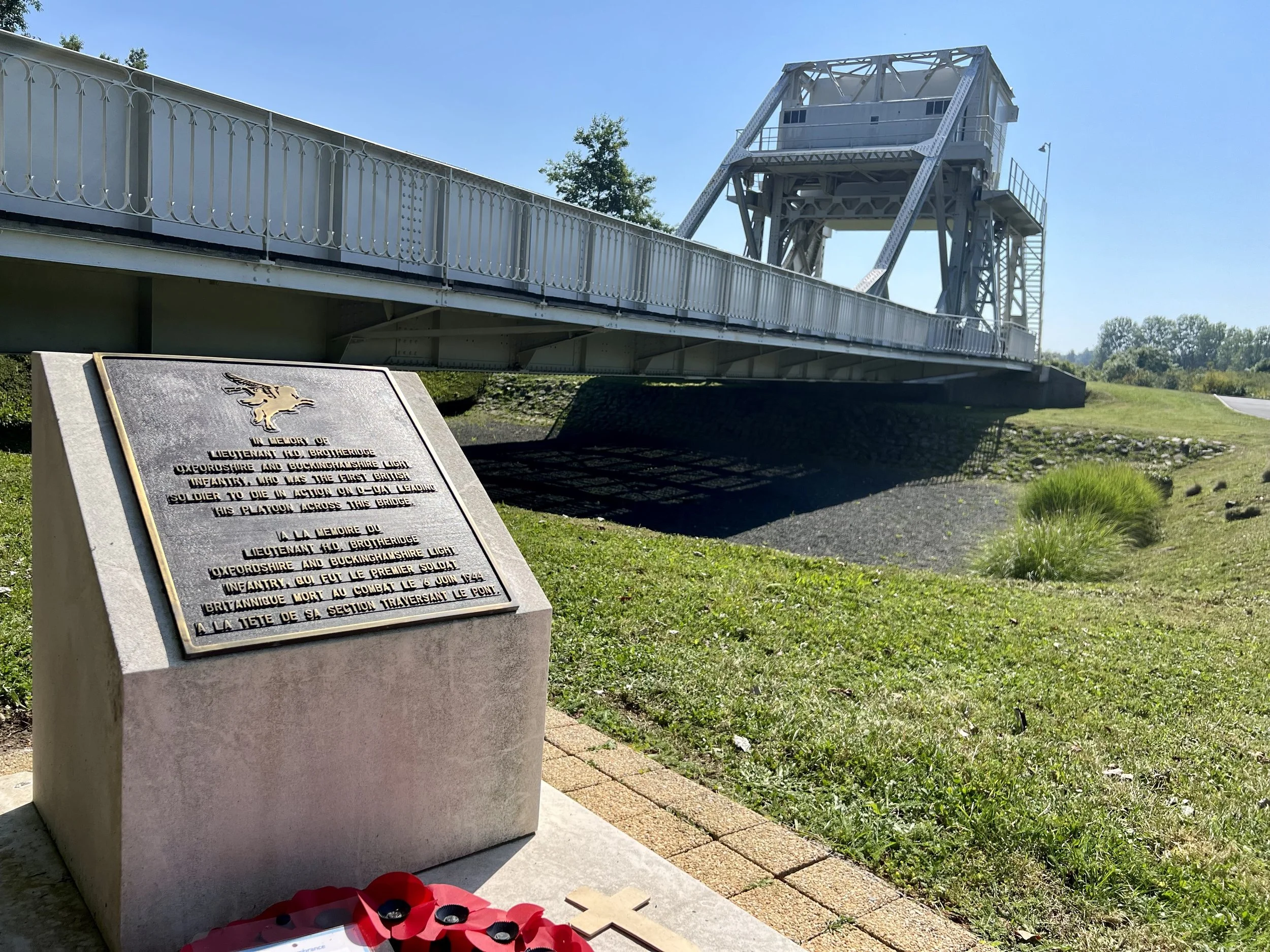 Memorial plaque in memory of Lieutenant Harold Brotherhood, a British soldier from Oxfordshire and Buckinghamshire Light Infantry, who died in action on June 6, 1944, crossing a bridge. The plaque is placed beside a bridge structure with a blue sky and green grass in the background.