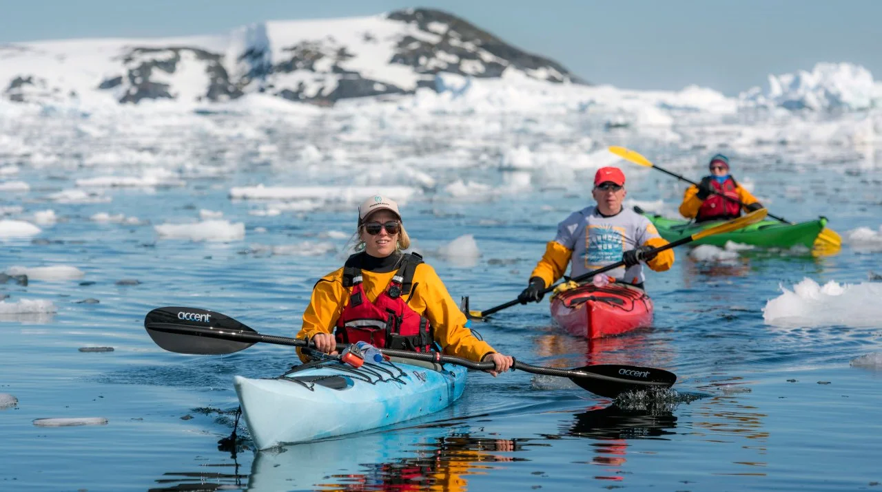 Three people kayaking in icy water with snow-covered mountains in the background.