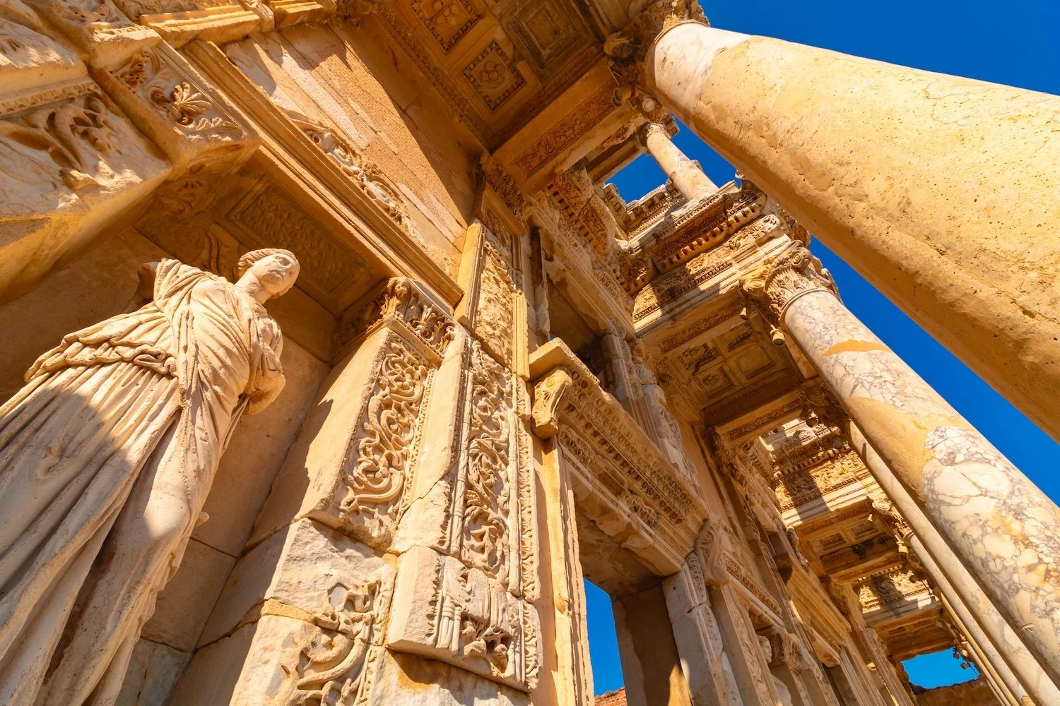Ancient stone ruins with carved columns, statues, and elaborate decorative friezes against a clear blue sky.