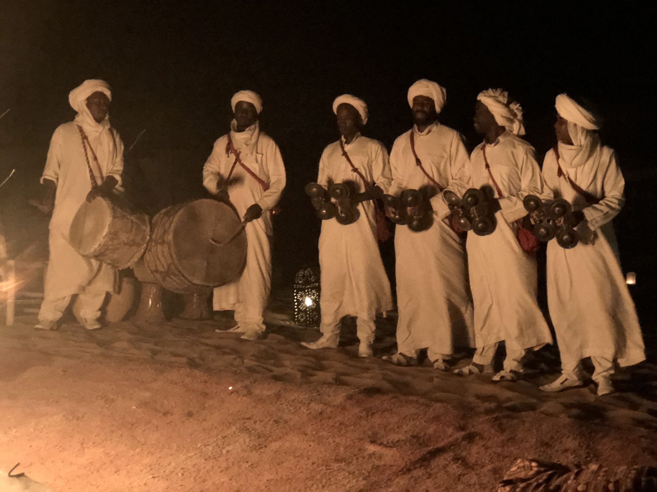 Group of six men dressed in traditional white clothing and head coverings, playing drums and percussion instruments at night.