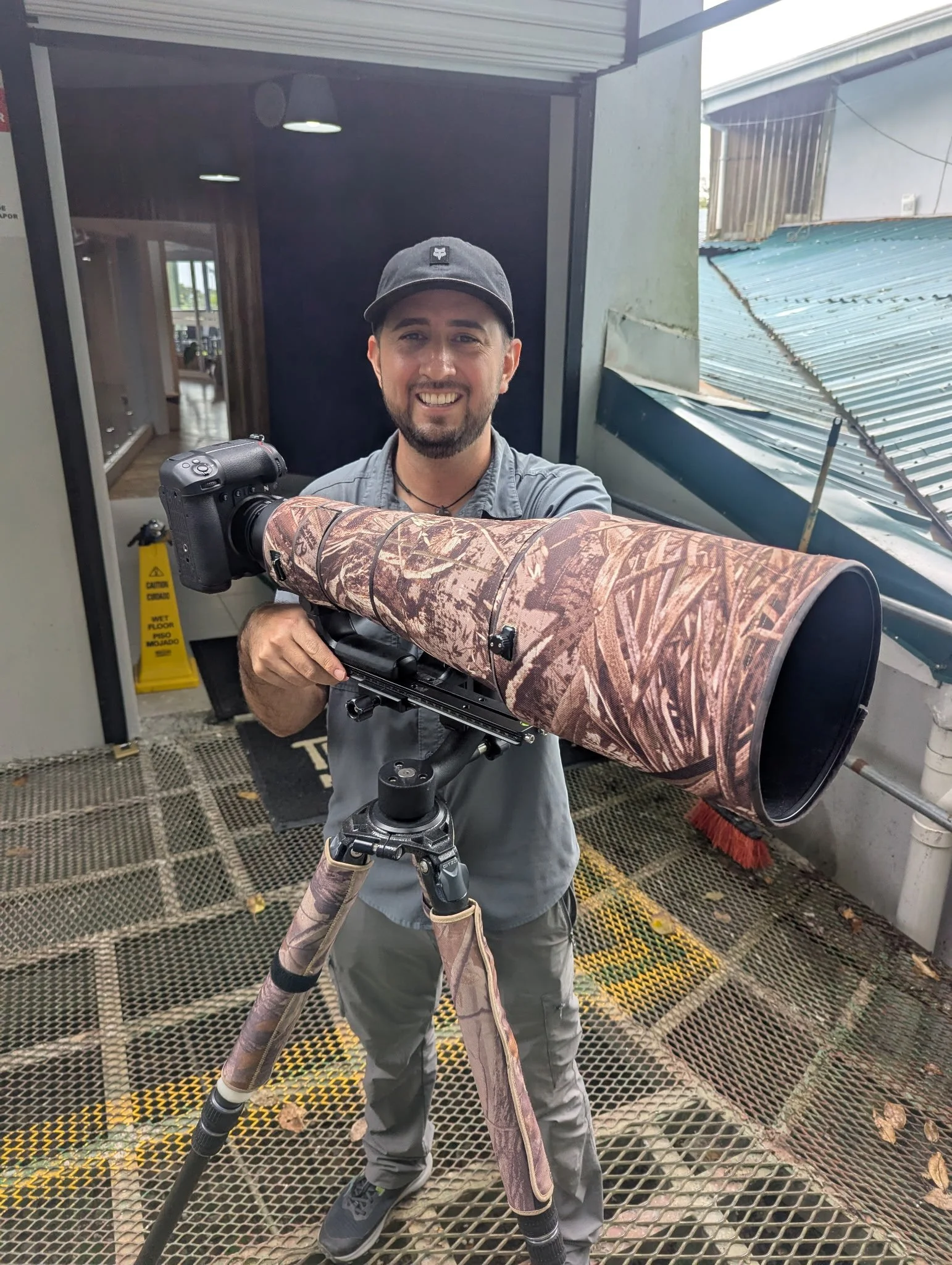 Man smiling at camera standing outside with a large spotting scope mounted on a tripod, dressed in gray shirt, pants, and a cap.