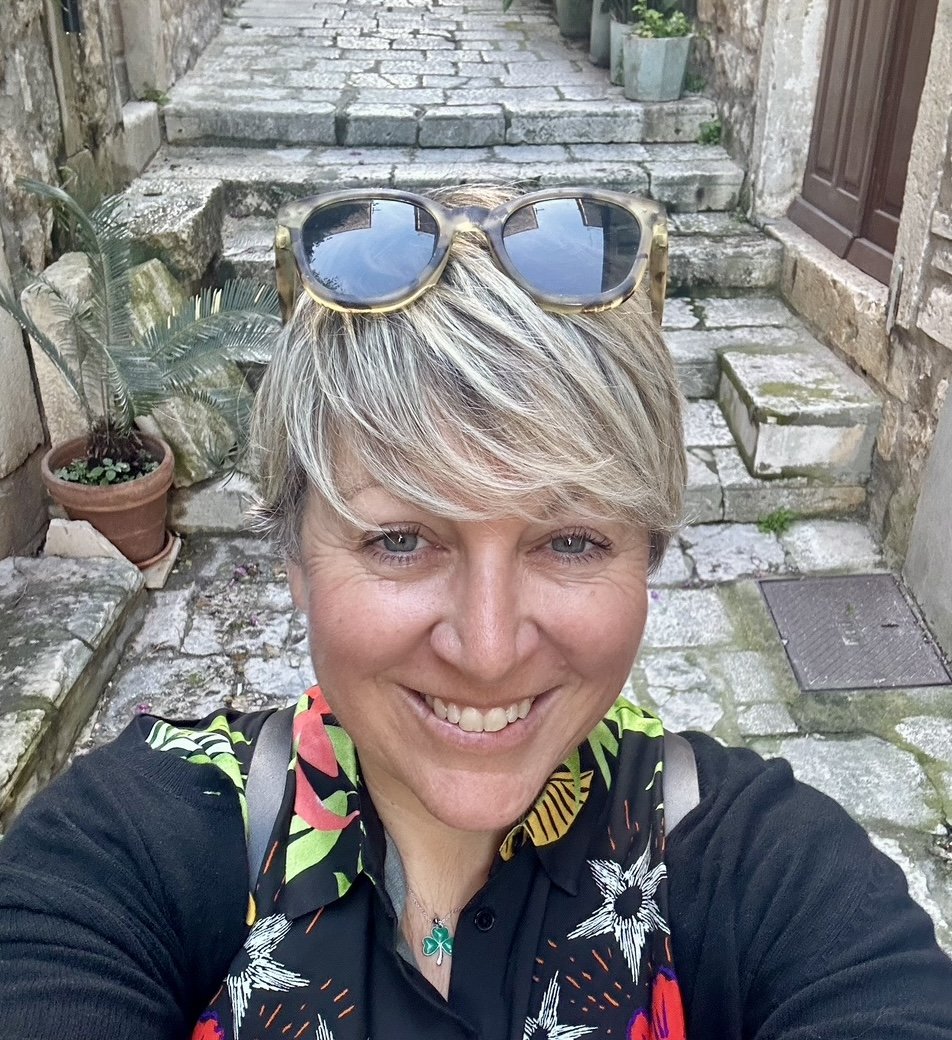 A woman with short blonde hair and sunglasses on her head taking a selfie outdoors on a stone staircase. She is wearing a black shirt with a colorful floral pattern and a necklace with a green pendant. There are potted plants nearby and a stone wall with a door in the background.