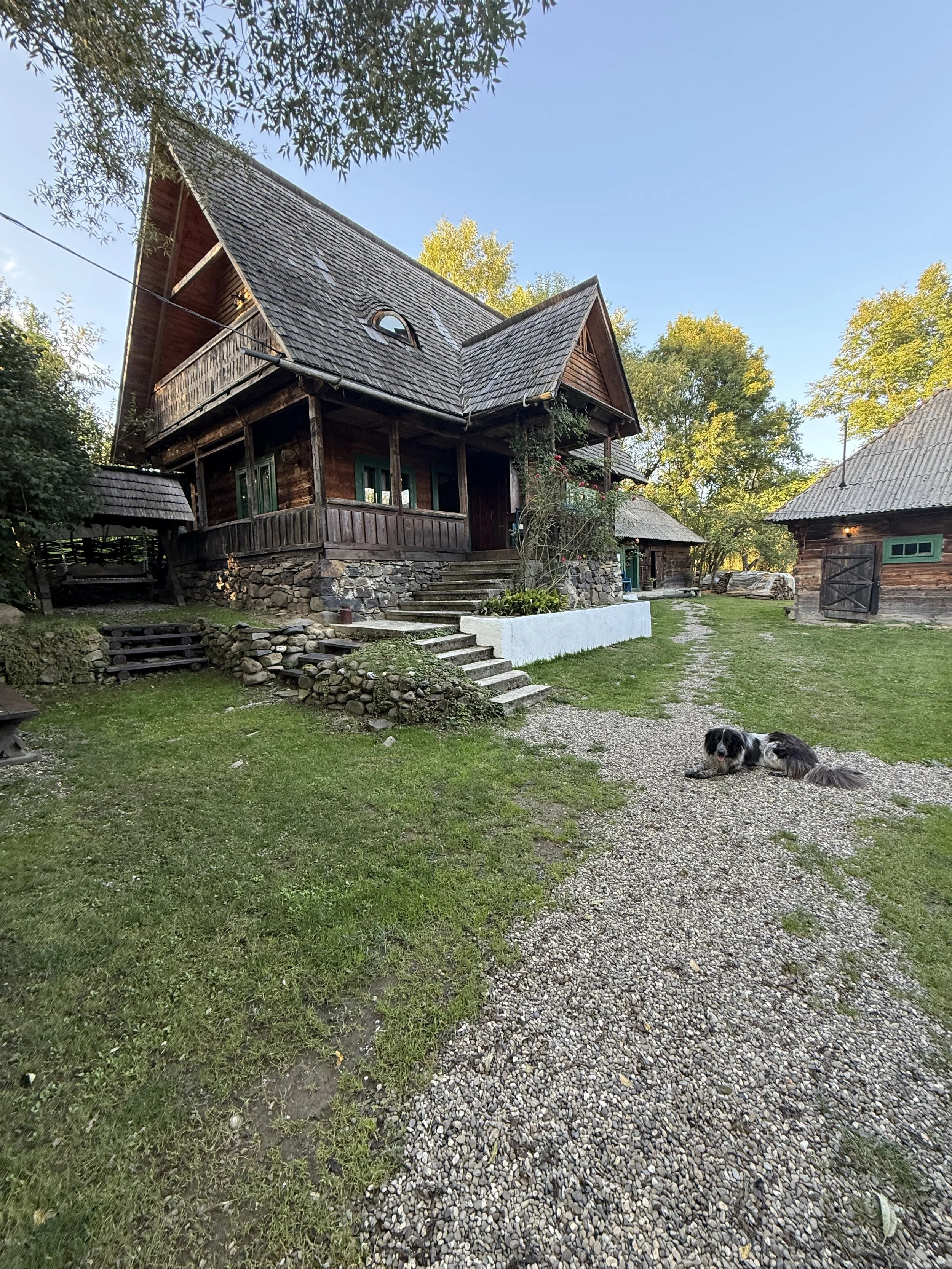 A rustic wooden house with a steep roof and stone foundation, surrounded by green grass and trees, with a gravel pathway and a dog lying on the ground.