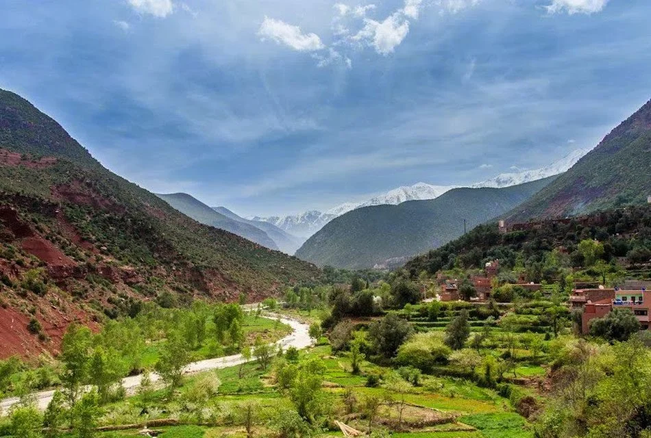 Scenic valley with green terraced agricultural fields, scattered houses, lush vegetation, and mountain ranges in the background under a partly cloudy sky.