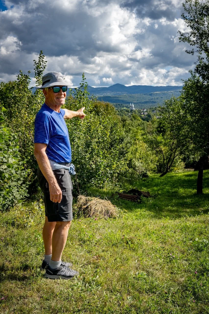 Older man wearing a blue shirt, gray shorts, a hat, and sunglasses, standing on grass in a green area, pointing toward distant mountains and a church steeple visible over the trees under a partly cloudy sky.