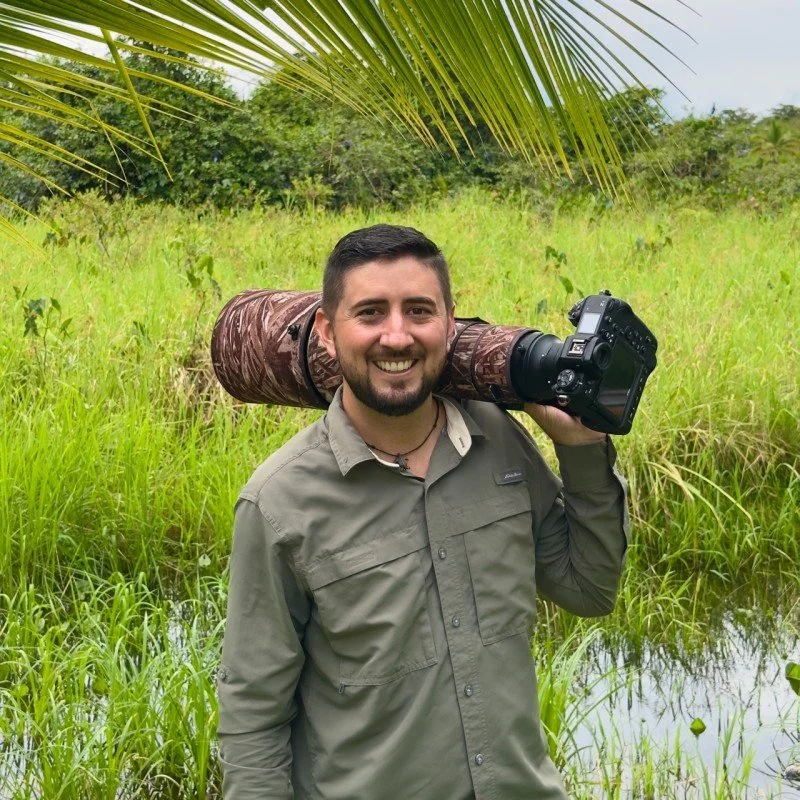 Man standing outdoors in a green, grassy area holding a camera with a large lens on his shoulder, smiling, with palm leaves overhead.