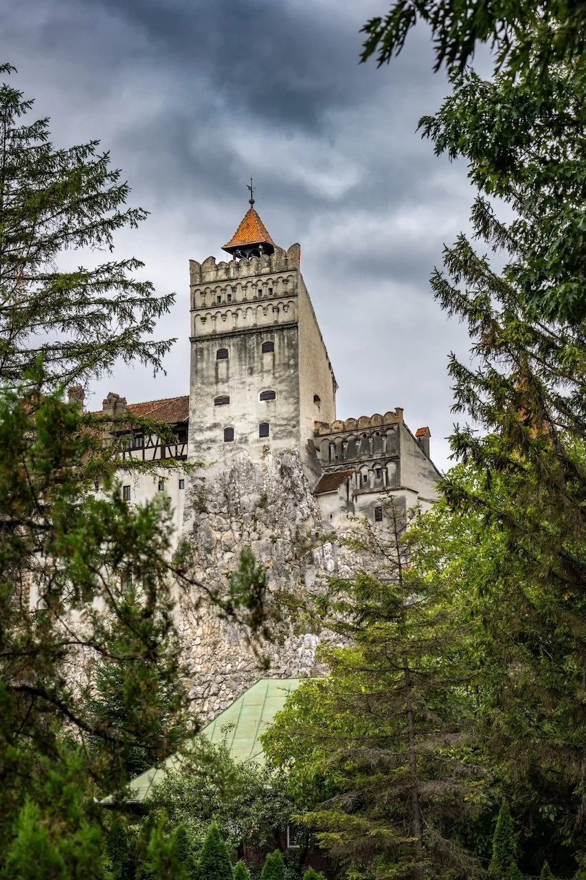 A medieval castle with a tall tower surrounded by green trees, with a cloudy sky overhead.
