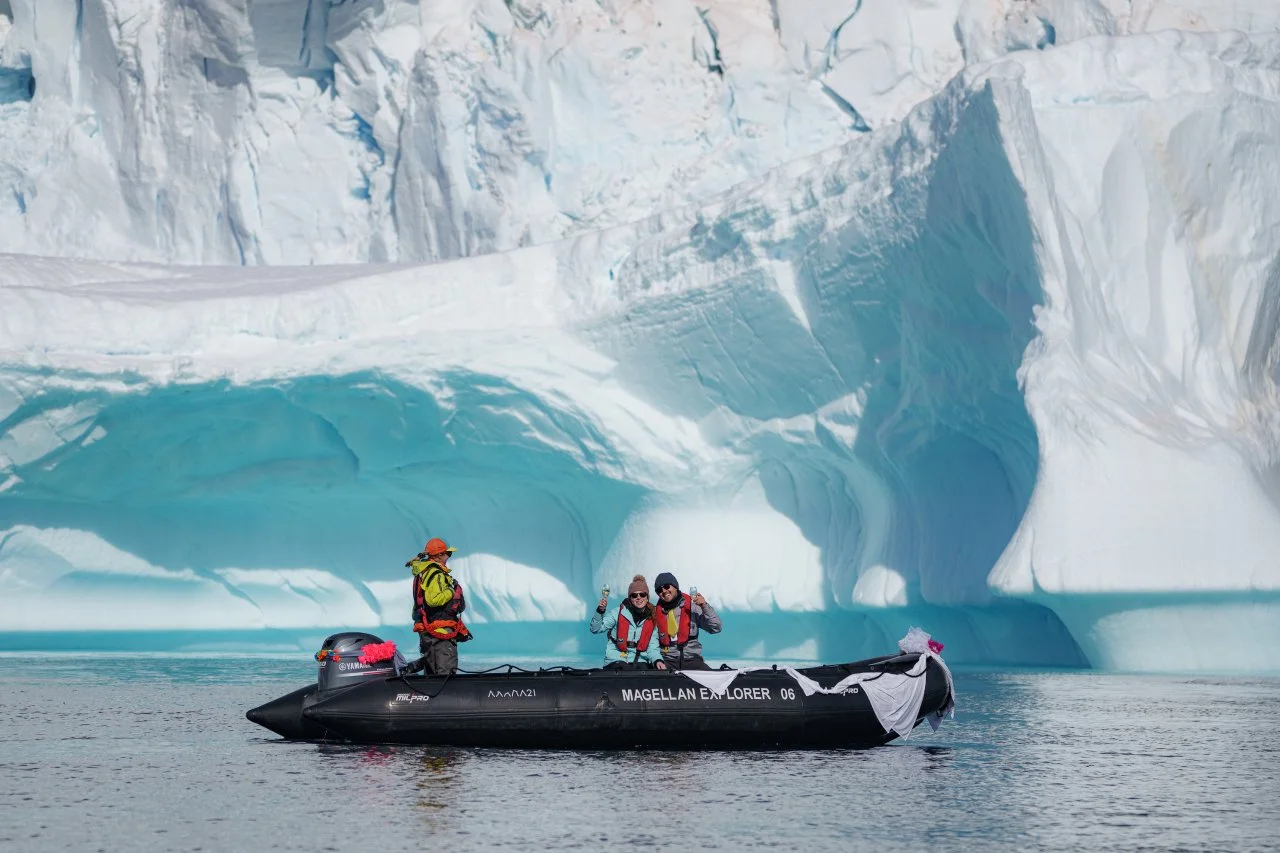 Three people in cold-weather gear on a black inflatable boat near an icy landscape with a large ice formation and glacier in the background.