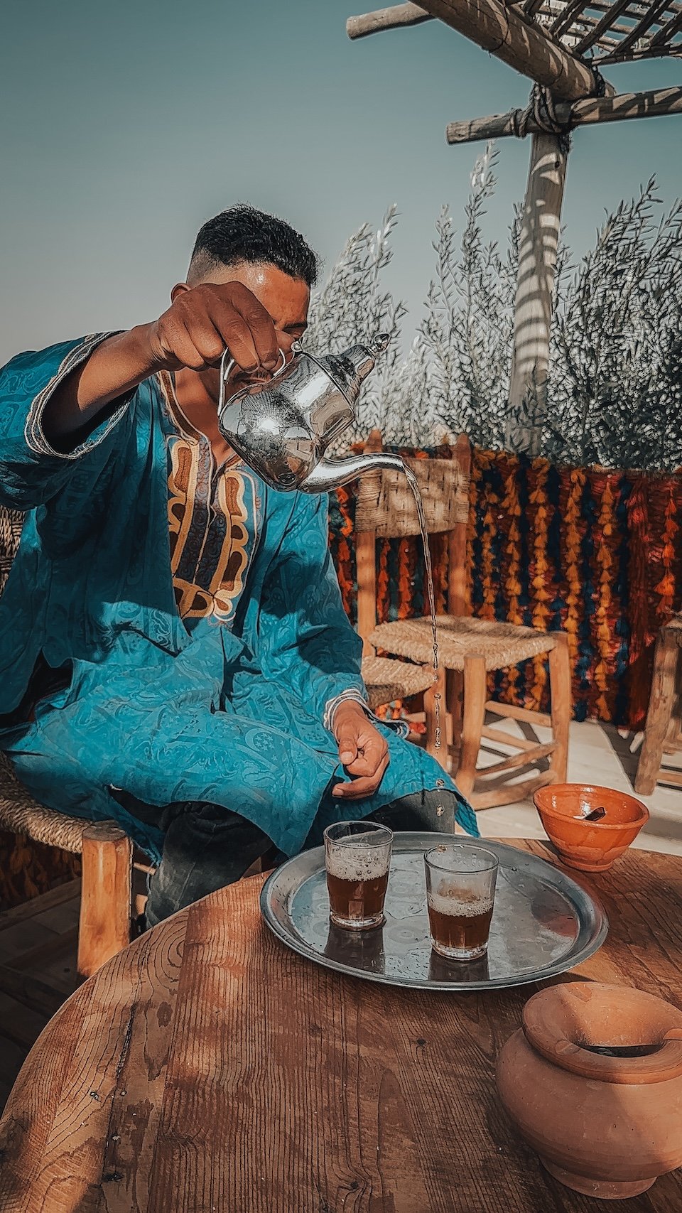 Person in traditional blue clothing pouring tea from a kettle into glasses on a tray, sitting at a round wooden table in a decorated outdoor setting.