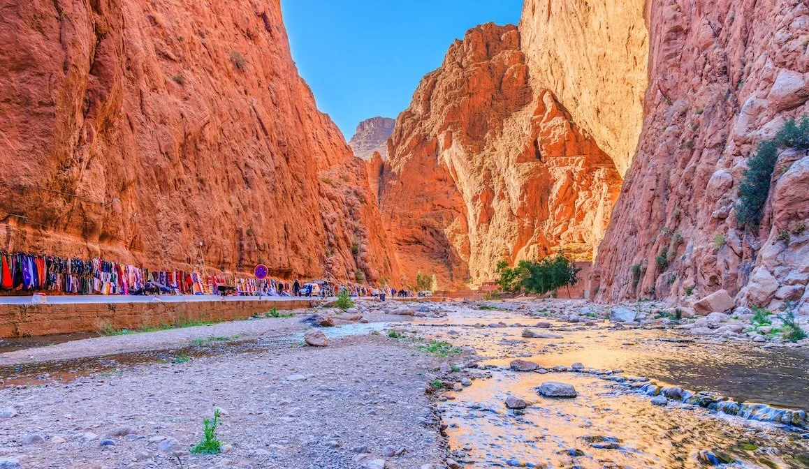 A narrow canyon with tall red and orange rock walls, a river flowing through the rocky valley, and a marketplace with colorful cloths along a pathway on the left side.