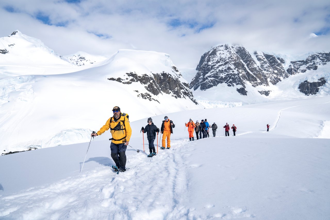 Group of people hiking in snowy mountain landscape with snow-covered peaks and cloudy sky.