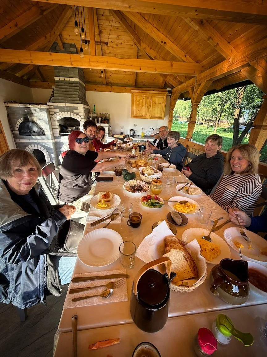 A group of people sitting around a long wooden table inside a cozy, wooden-ceiling pavilion enjoying a meal together. The table has plates of food, bread, and drinks, with a fireplace in the background and greenery visible outside through the open windows.