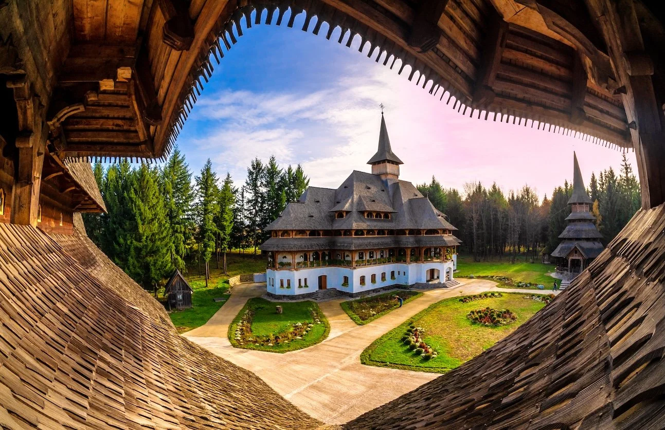 View of a traditional wooden church with a garden, seen through a decorative wooden frame or window, surrounded by trees and a partly cloudy sky.