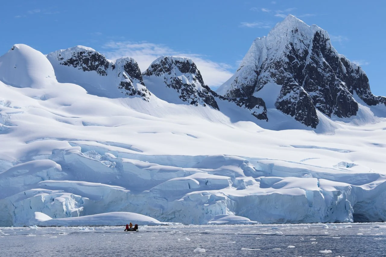 A snowy mountain landscape with large ice formations and two people in a small boat on the water in the foreground.