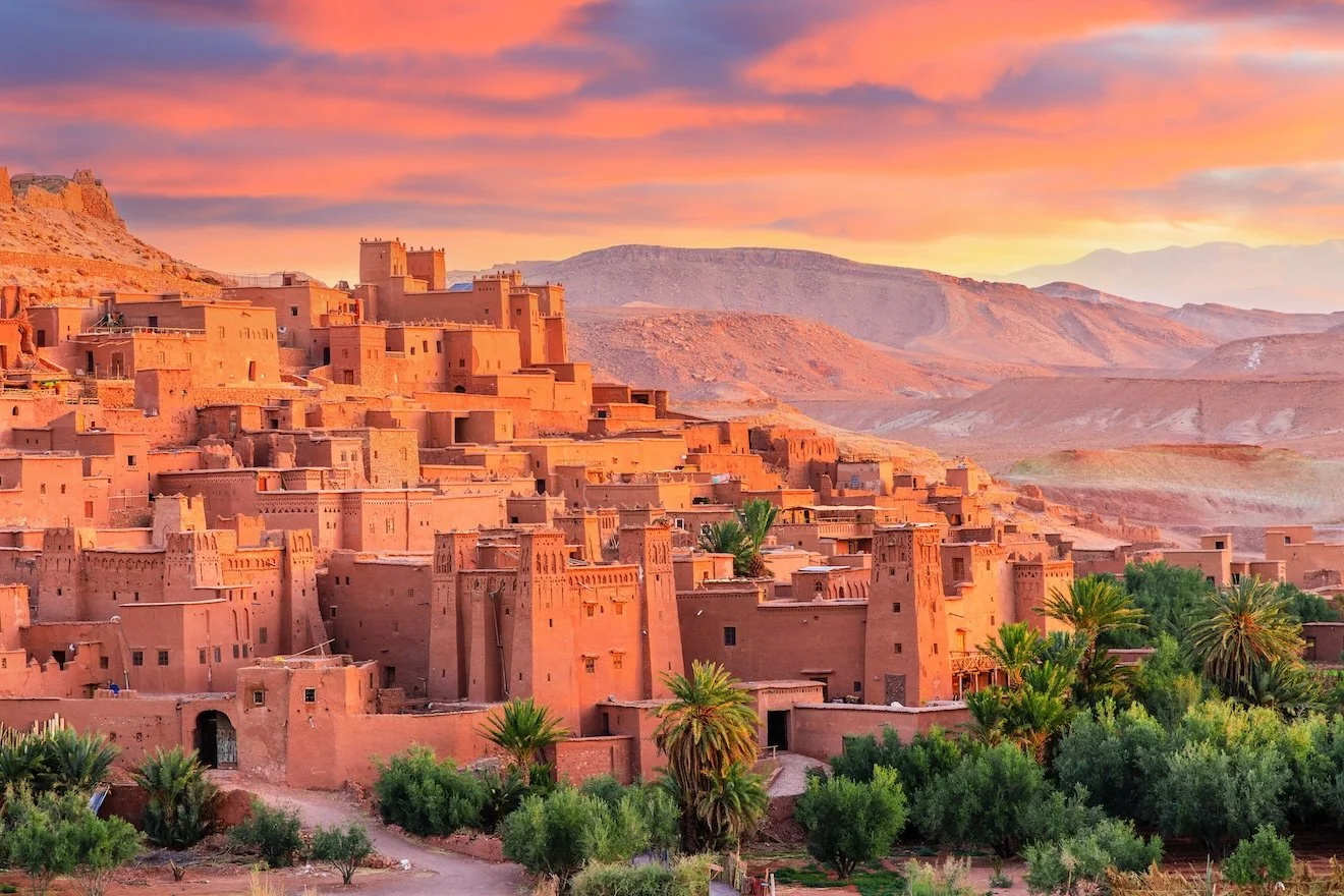 Sunset over a traditional desert city with adobe houses, surrounded by palm trees and mountains in the background.