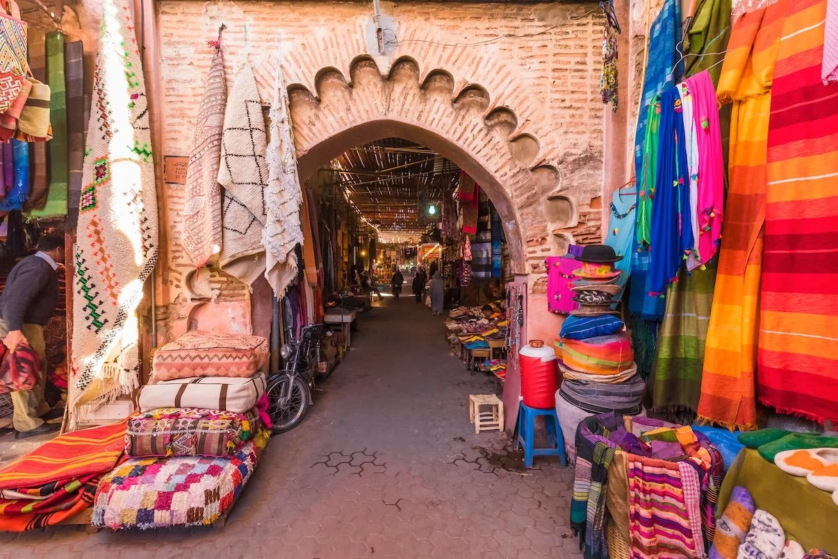 A market alleyway with colorful textiles and rugs hanging on both sides, and an archway made of brick with decorative scalloped edges leading into the market.