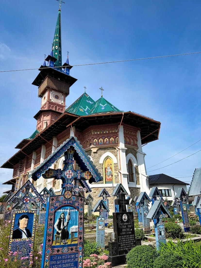 Colorful church with green and gold rooftops and multiple steeples, surrounded by a cemetery with decorated graves, some with painted images and Christian symbols, under a clear blue sky.