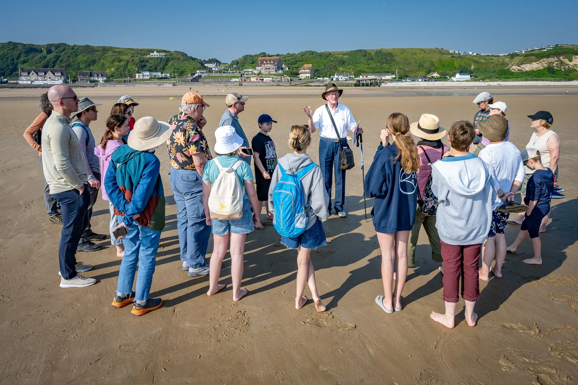 A group of people on a beach listening to a guide, with some wearing hats and backpacks, standing in a semi-circle on sand, with houses and green hills in the background.