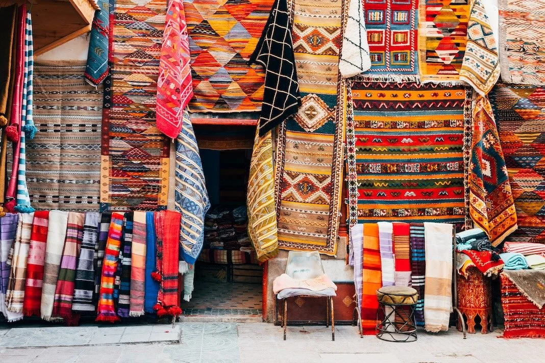A display of colorful woven and embroidered textiles, including rugs, scarves, and throws, arranged outside a shop.