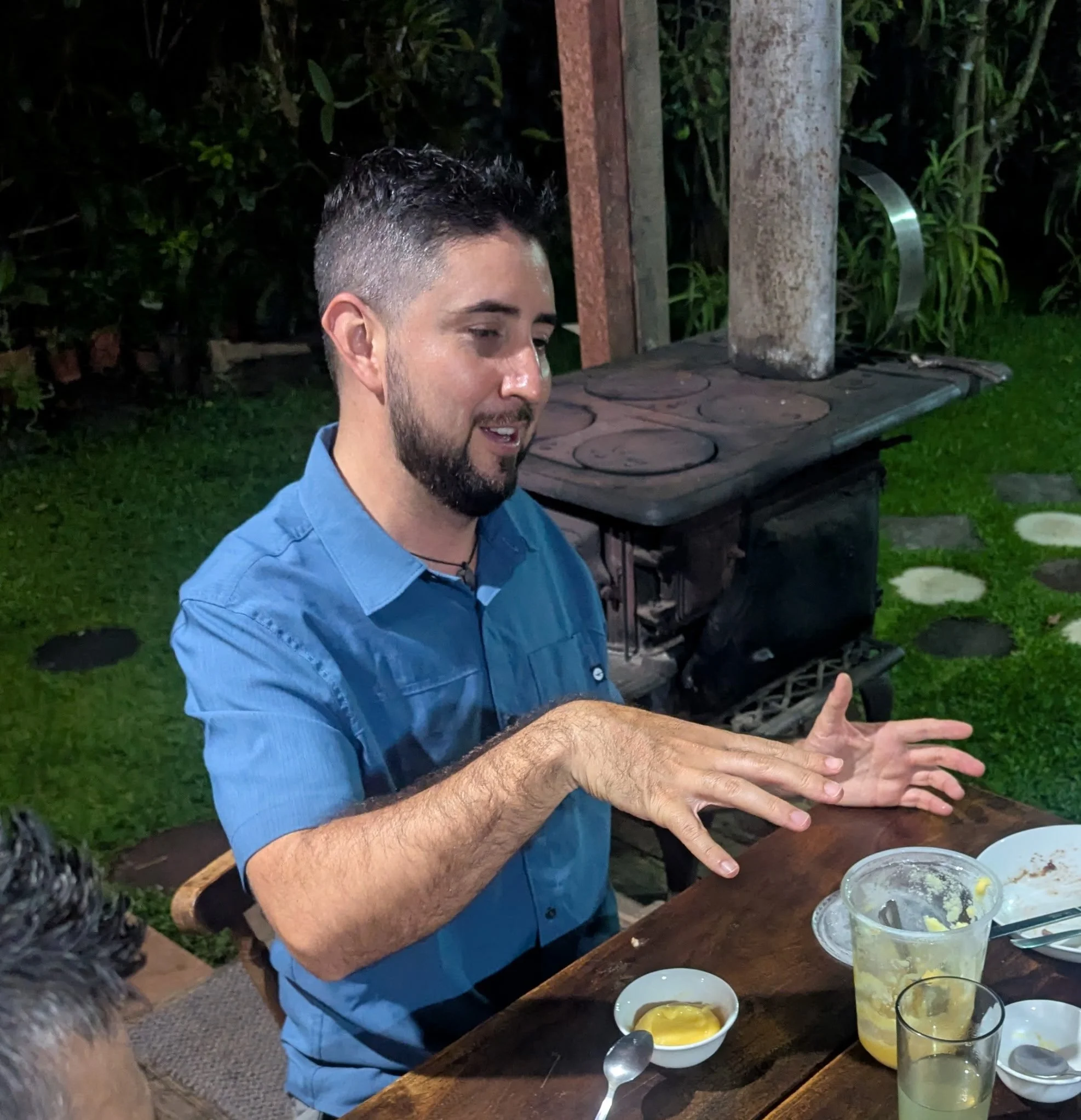 A man in a blue shirt sitting at a wooden table outdoors, gesturing with his hands while talking, with dishes and drinks on the table, and a wood stove in the background.
