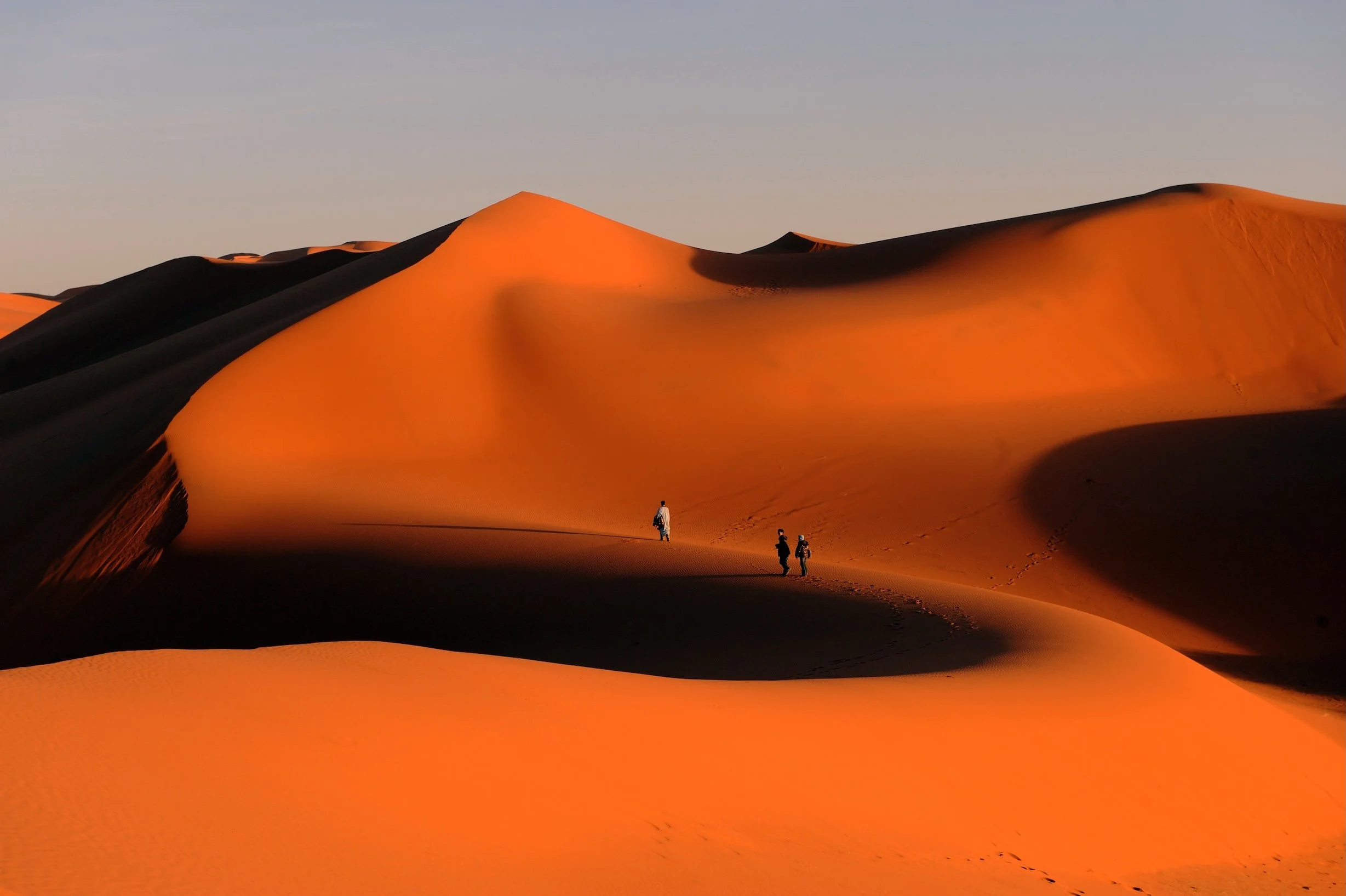 Three people walking through vast orange sand dunes in a desert during sunset.