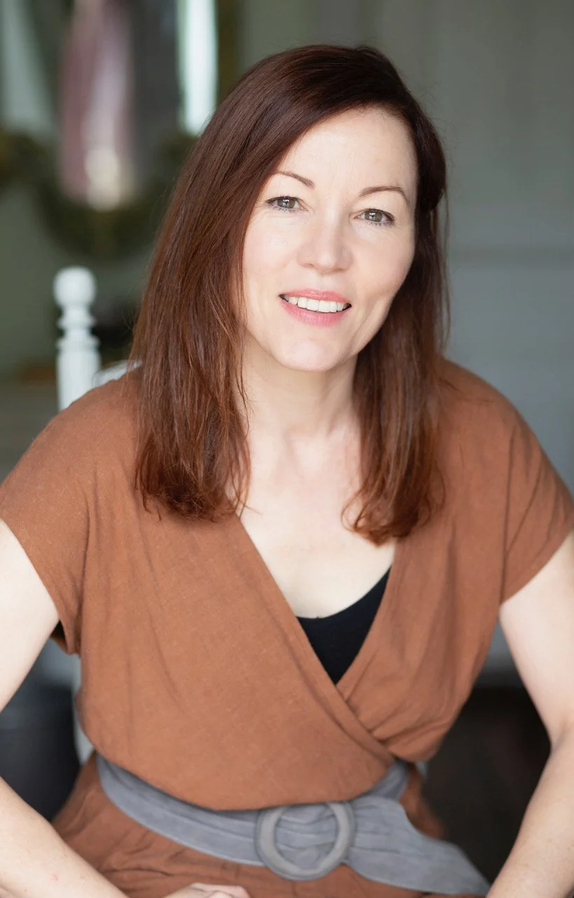A woman with shoulder-length reddish-brown hair, wearing a brown top, smiling at the camera. She is seated indoors with a blurred background.