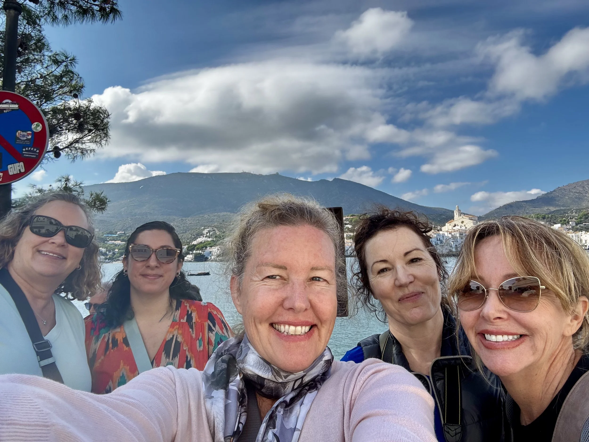 Group of five women taking a selfie by a river, with mountains and a cloudy sky in the background.