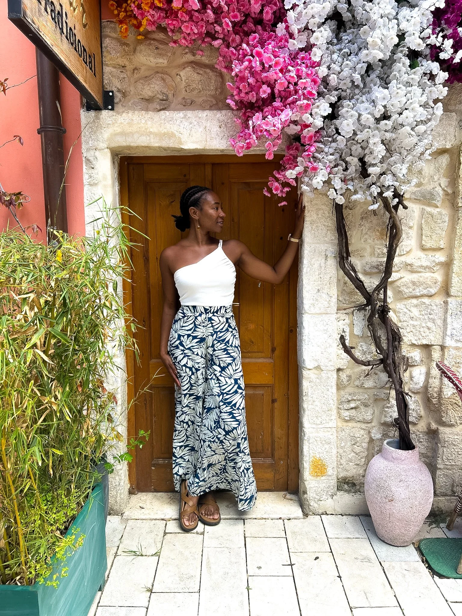 Woman in a white top and patterned maxi skirt posing in a stone doorway framed by pink and white flowers, Vlore, Albania
