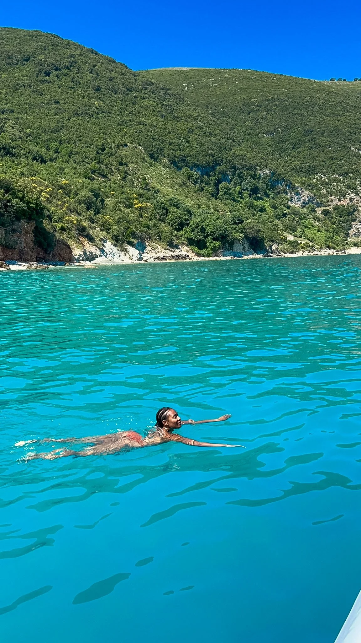 Woman swimming in clear turquoise water below green hills on the Karaburun Peninsula, Albania