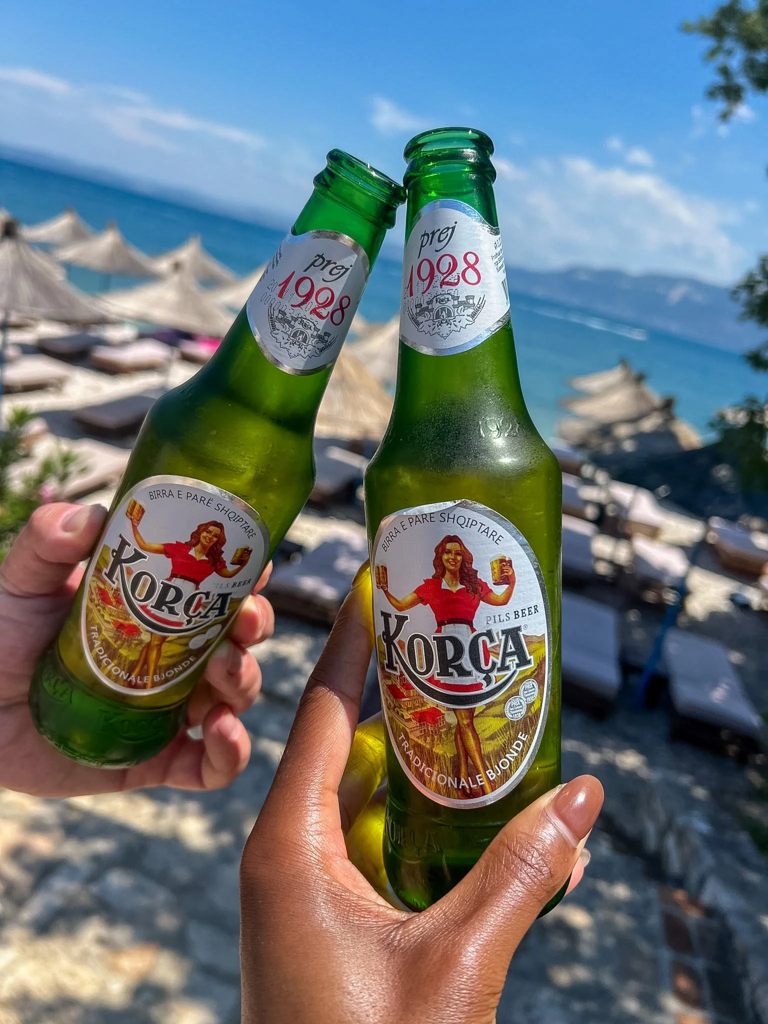 Two hands raising bottles of Korça beer over a beach club, Vlore, Albania