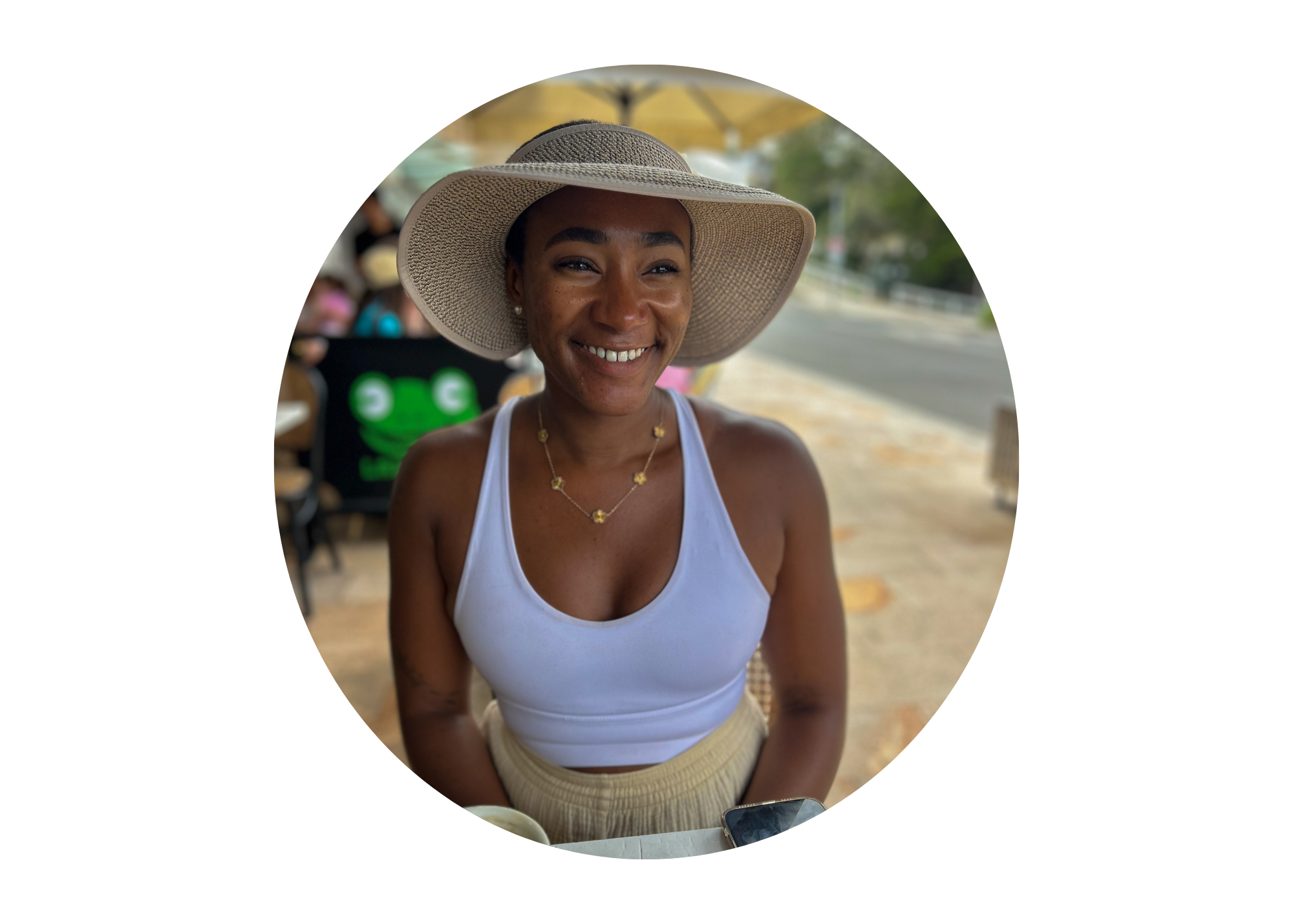 Smiling woman wearing a wide-brimmed hat, white tank top, and gold jewelry, sitting outdoors at a table