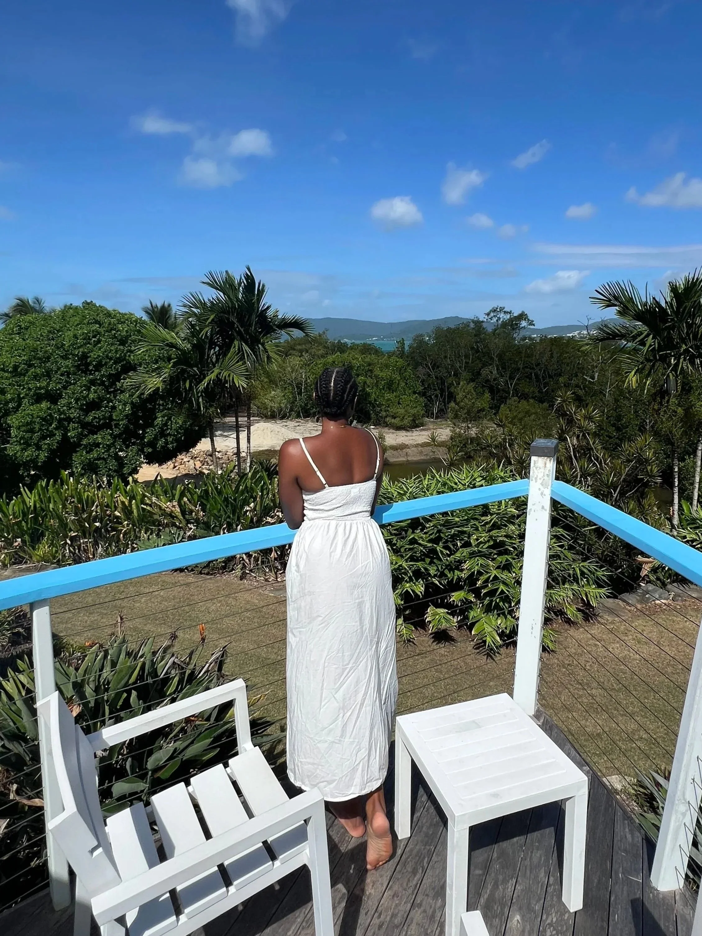 A woman in a white dress standing barefoot on a balcony, looking at lush green trees, water, and distant hills under a partly cloudy blue sky.