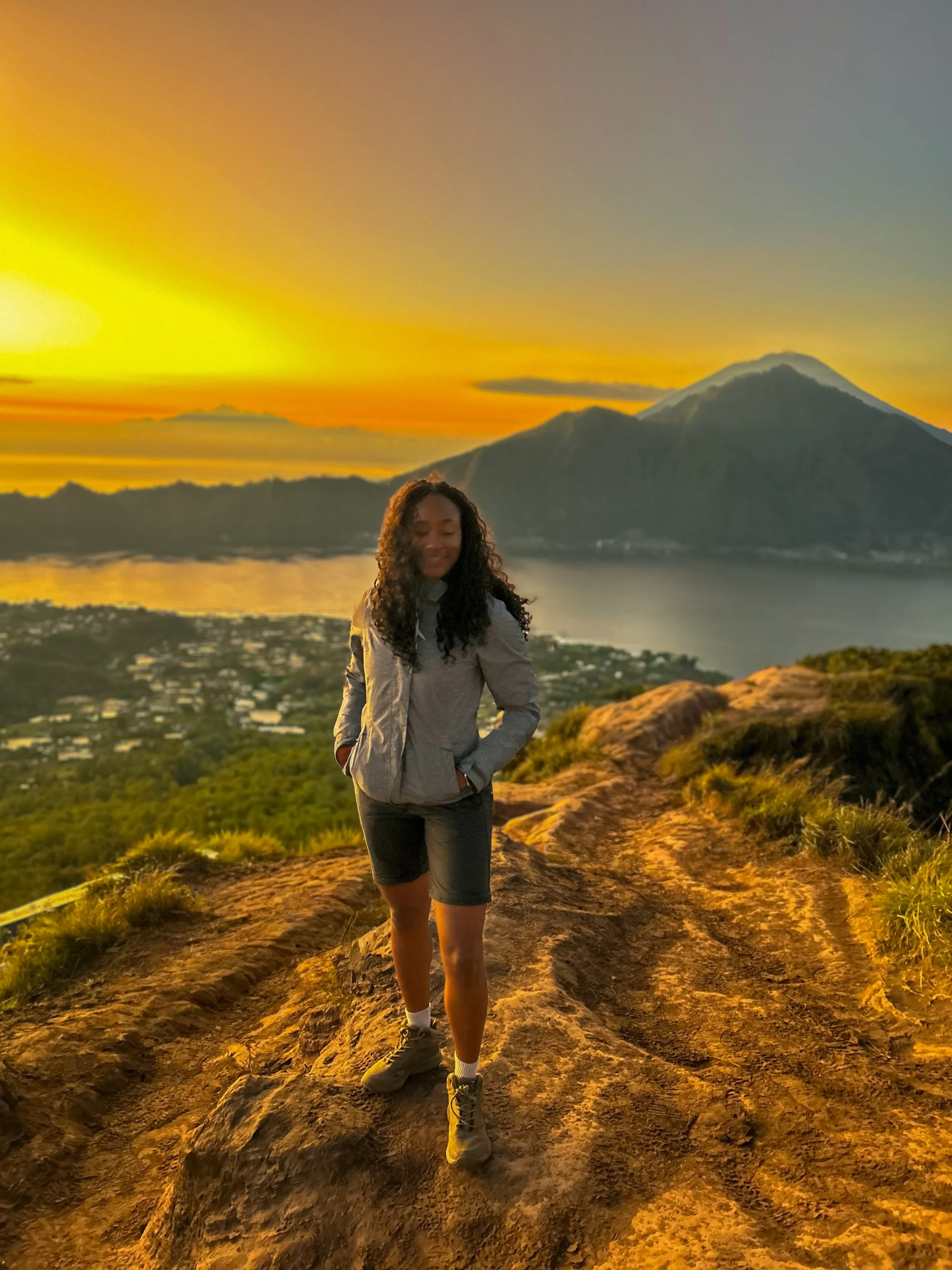 A woman standing on a rocky trail during sunset, with a large mountain and lake in the background.